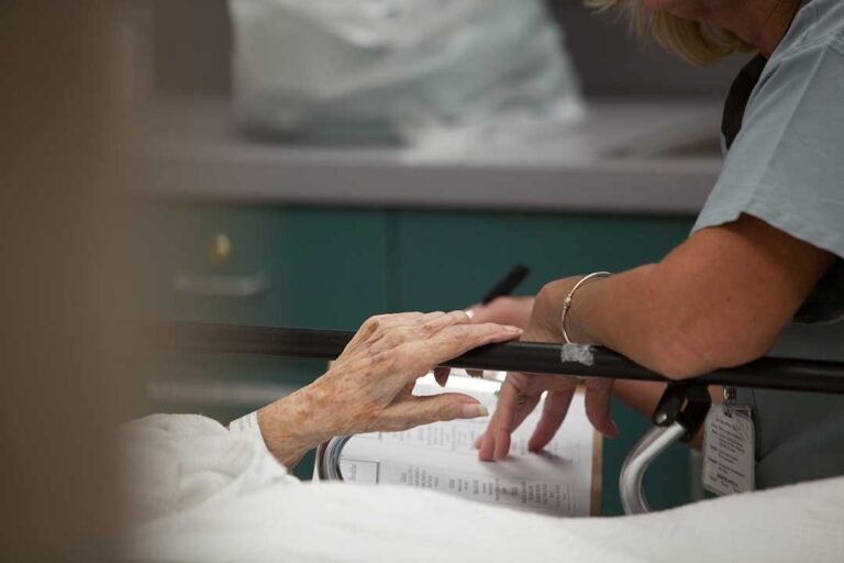 Two older adults talk at a kitchen table with a concerned family member, papers and medication bottles nearby.